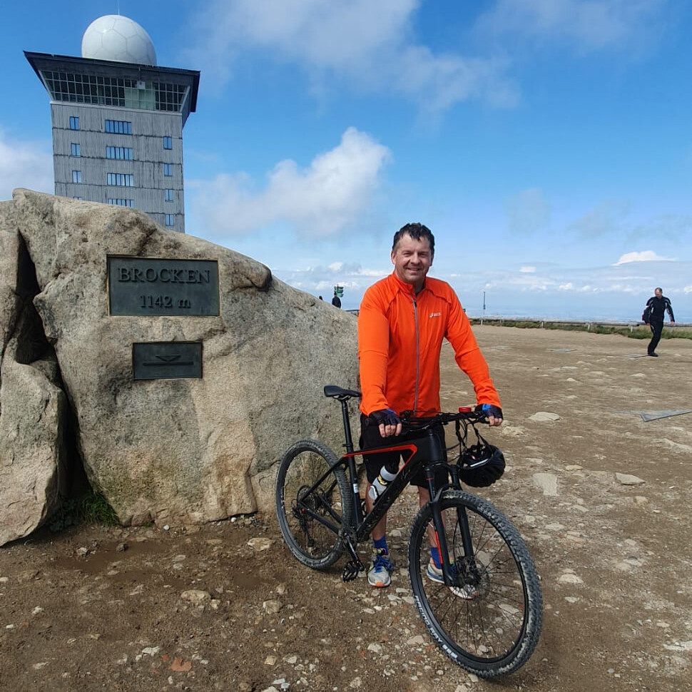 Sven Schulze steht bei blauem Himmel in orangener Jacke mit seinem Fahrrad am höchsten Punkt des Brocken